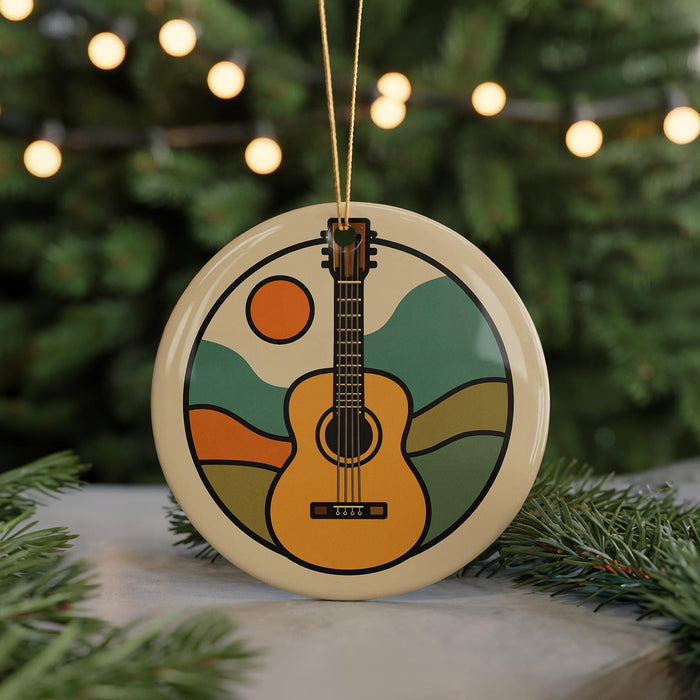 A round ornament featuring a guitar with a sun and mountain landscape design, hanging from a string against a blurred background of a Christmas tree with lights.