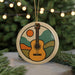 A round ornament featuring a guitar with a sun and mountain landscape design, hanging from a string against a blurred background of a Christmas tree with lights.
