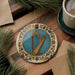 A decorative ornament featuring a harp is displayed on a wooden surface, surrounded by pine branches and a cup of coffee.
