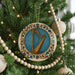 A round ornament with a harp design is hanging on a Christmas tree, surrounded by other ornaments and lights.