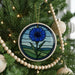 A blue and green stained glass ornament with a flower design hangs on a Christmas tree, surrounded by white beads and greenery.
