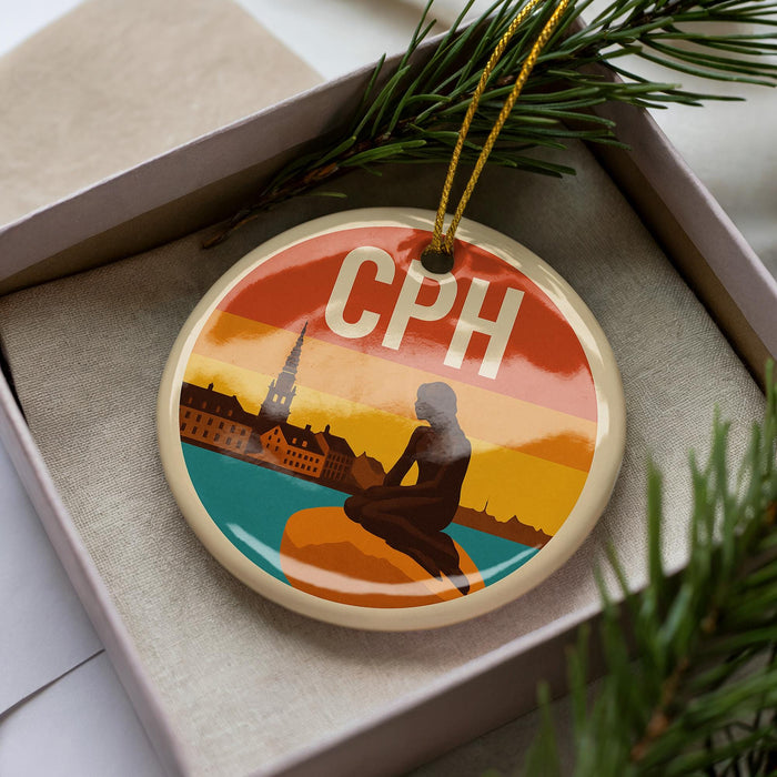 A round ornament with a silhouette of a person sitting on a rock, surrounded by a colorful sunset scene, is displayed in a box with pine branches.