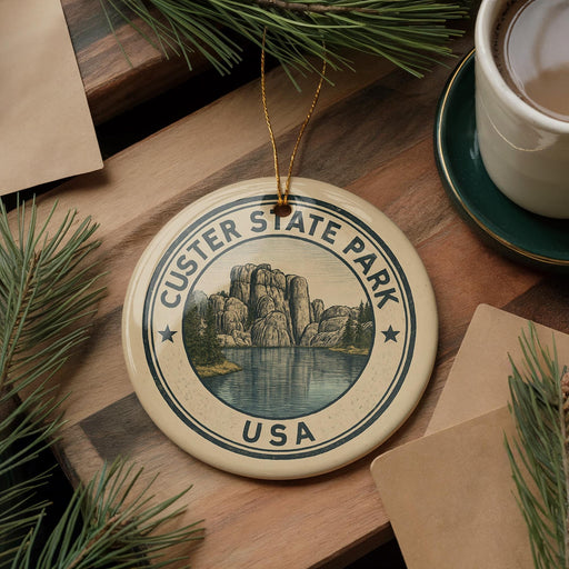 A ceramic ornament with the text "CUSTER STATE PARK USA" and an illustration of a mountain landscape is displayed on a wooden surface, accompanied by pine branches and a cup of coffee.