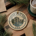 A ceramic ornament with the text "CUSTER STATE PARK USA" and an illustration of a mountain landscape is displayed on a wooden surface, accompanied by pine branches and a cup of coffee.