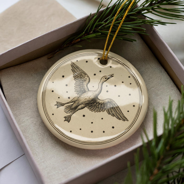 A ceramic ornament in the shape of a bird, with a starry background, is displayed in a box with pine branches.