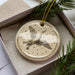A ceramic ornament in the shape of a bird, with a starry background, is displayed in a box with pine branches.