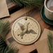 a round ornament with a bird illustration, placed on a wooden surface next to a cup of coffee and some pine branches.