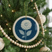 A blue and white ceramic ornament with a white flower design hangs from a Christmas tree, surrounded by white wooden beads and twinkling lights.