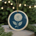 A blue and white ceramic ornament featuring a dahlia flower is displayed in the center of the image, surrounded by pine branches and twinkling lights.