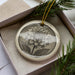A decorative ceramic ornament featuring a detailed floral design is displayed in a box, surrounded by pine branches.