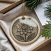 A decorative ornament featuring a detailed illustration of a flower is displayed in a box, surrounded by pine branches and greenery.