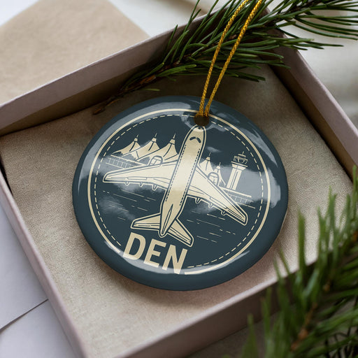 A circular ornament with a picture of an airplane and the word "DEN" on it, placed in a box with pine branches.