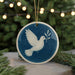 A blue ceramic ornament with a white dove in flight and a peace sign, hanging from a string.