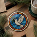 A stained glass ornament featuring a white dove in flight is displayed on a wooden surface, surrounded by pine branches and a cup of coffee.