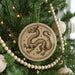 A round ornament featuring a dragon design is hanging on a Christmas tree, surrounded by other ornaments and lights.