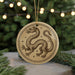 A circular ornament featuring a detailed illustration of a dragon, with a starry background, is displayed in front of a blurred Christmas tree.