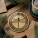 a decorative ceramic ornament with a dragonfly design, placed on a wooden surface alongside a cup of coffee and some pine branches.