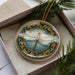 A decorative ceramic ornament in the shape of a dragonfly, with a blue and gold border, is displayed in a box surrounded by pine branches.