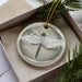 A ceramic ornament in the shape of a dragonfly, with a detailed illustration of the insect, is displayed in a box surrounded by pine branches.