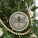 A ceramic ornament featuring a detailed illustration of a dragonfly is hanging from a Christmas tree, surrounded by ornaments and lights.