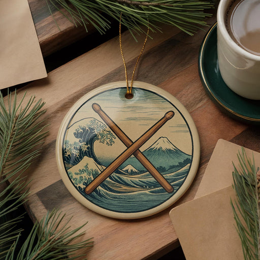 A circular ornament featuring a wave and mountain landscape with crossed drumsticks, placed on a wooden surface alongside a cup of coffee and some pine branches.