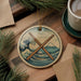 A circular ornament featuring a wave and mountain landscape with crossed drumsticks, placed on a wooden surface alongside a cup of coffee and some pine branches.