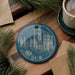 A blue ornament with a picture of a castle or cathedral, featuring the word "DUR" on it, is displayed on a wooden surface alongside a cup of coffee and some pine branches.