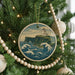 A Christmas tree decorated with a round ornament featuring a lighthouse on a cliff overlooking the ocean, surrounded by a string of beads.