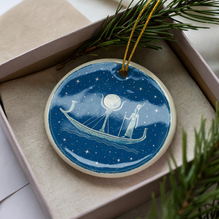 A blue and white ornament with a boat and a moon on a starry night scene, displayed in a box with pine branches.