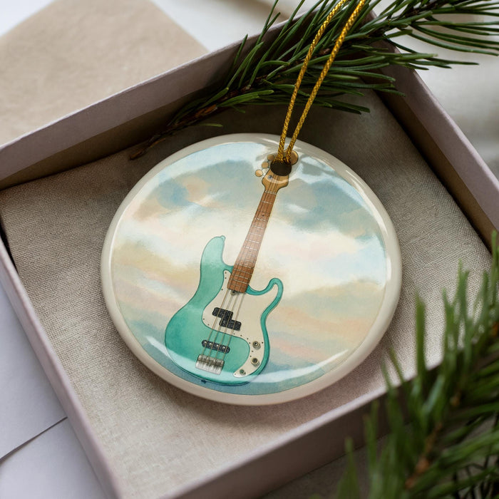 A ceramic ornament in the shape of a guitar, with a light blue and white color scheme, is displayed in a box with pine branches.