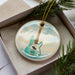 A ceramic ornament in the shape of a guitar, with a light blue and white color scheme, is displayed in a box with pine branches.