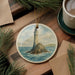 A round ceramic ornament depicting a lighthouse on a rocky island, surrounded by water, is displayed on a wooden surface alongside a cup of coffee and some pine branches.