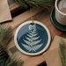 a blue and white circular ornament with a fern design, placed on a wooden surface alongside a cup of coffee and some pine branches.