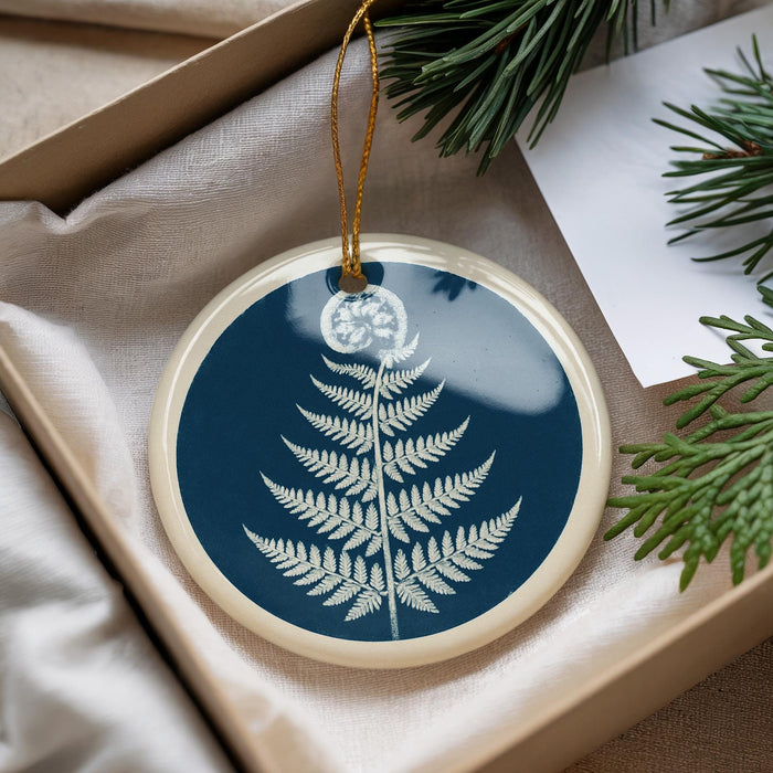 A blue and white ornament with a fern design is displayed in a box, surrounded by pine branches and a white card.