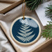 A blue and white ornament with a fern design is displayed in a box, surrounded by pine branches and a white card.