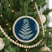A blue and white circular ornament with a fern design hangs from a Christmas tree, surrounded by white wooden beads and twinkling lights.