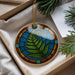 A decorative ornament with a fern leaf design is displayed in a box, surrounded by pine branches and a white card.