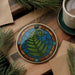 A decorative ornament featuring a fern leaf design is displayed on a wooden surface, accompanied by a cup of coffee and some pine branches.