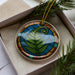 A decorative ornament in a box, featuring a colorful design of a fern leaf and a stained glass motif.