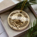 A decorative ornament in a box, featuring a fire lookout tower and crossed axes, surrounded by pine branches.