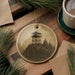 A round ornament featuring a lighthouse on a mountain, surrounded by pine branches and a cup of coffee on a wooden surface.
