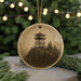 A round ornament featuring a black and white image of a tower on a mountain, surrounded by pine trees, with a string of lights in the background.