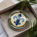 A decorative ornament in a box, featuring a blue fleur-de-lis design with gold and blue accents.