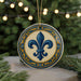 A decorative ornament featuring a blue fleur-de-lis symbol is displayed on a table, surrounded by pine branches and twinkling lights.