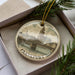 A decorative ornament featuring a cityscape with a bridge, displayed in a box with pine branches.