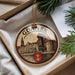 A circular ornament with a drawing of a castle and a town, surrounded by a border, is displayed in a box with pine branches.