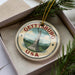 A decorative ornament in a box, featuring a circular design with the text "GETTYSBURG USA" and an illustration of a statue.