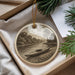 A decorative ornament featuring a canoe on a river, surrounded by trees and a cloudy sky, is displayed in a box with pine branches.