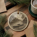 a round ceramic ornament with a black and white illustration of a canoe on a river, surrounded by pine branches and a cup of coffee on a wooden surface.