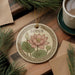 A decorative ceramic ornament featuring a pink lotus flower and green leaves, with the word "LOTUS" written on it, is displayed on a wooden surface alongside a cup of coffee and some pine branches.